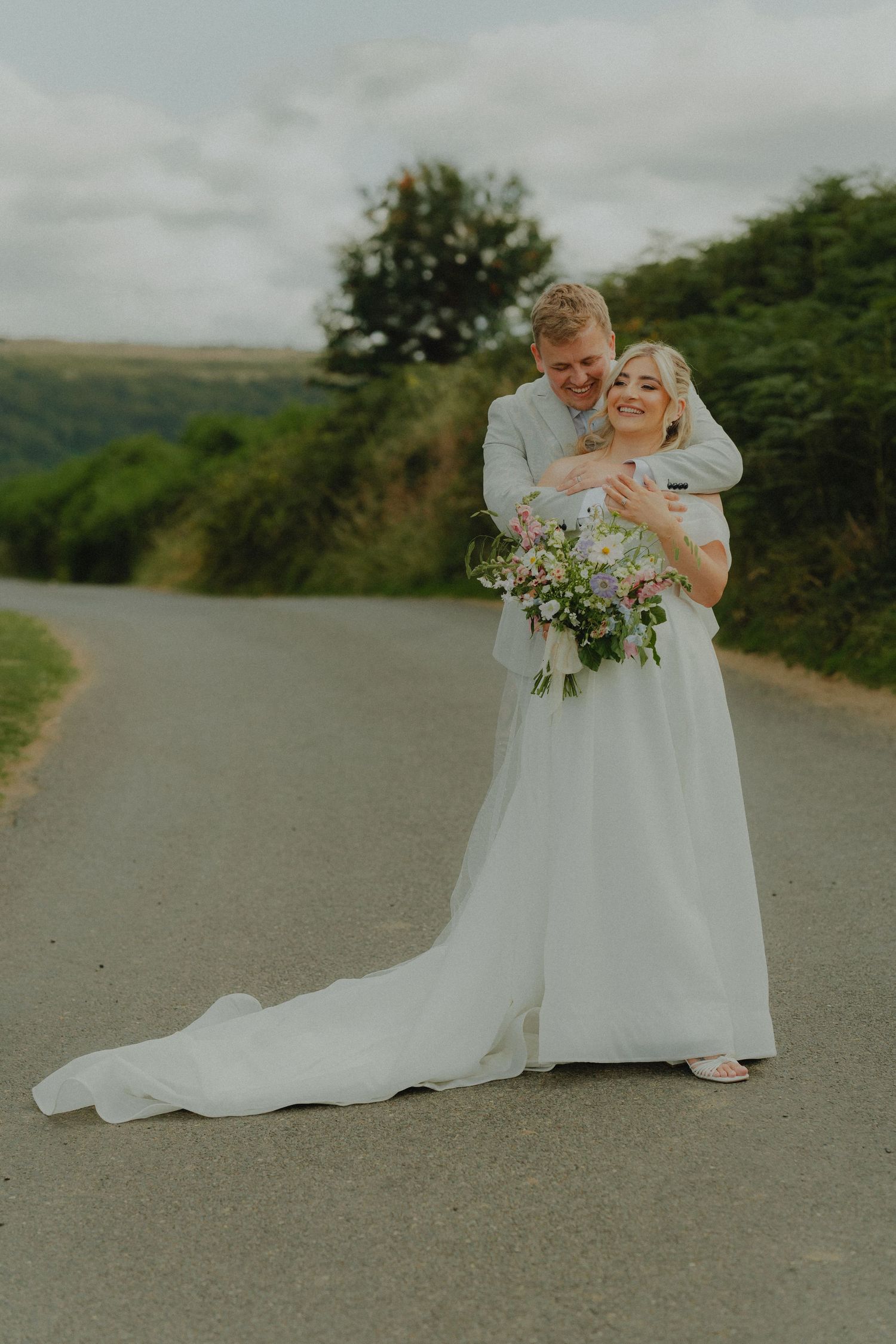 A groom embraces his smiling bride in a white gown holding a floral bouquet on a scenic countryside road.