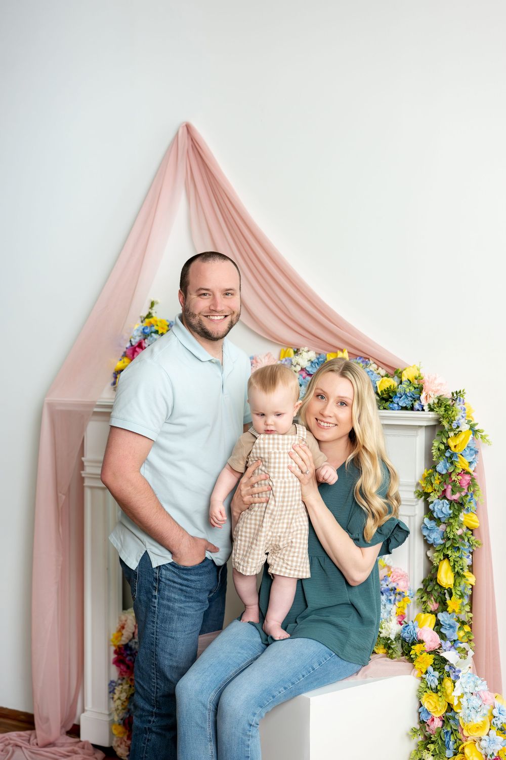 Smiling family portrait with father, blonde mother, and baby near spring floral fireplace backdrop.