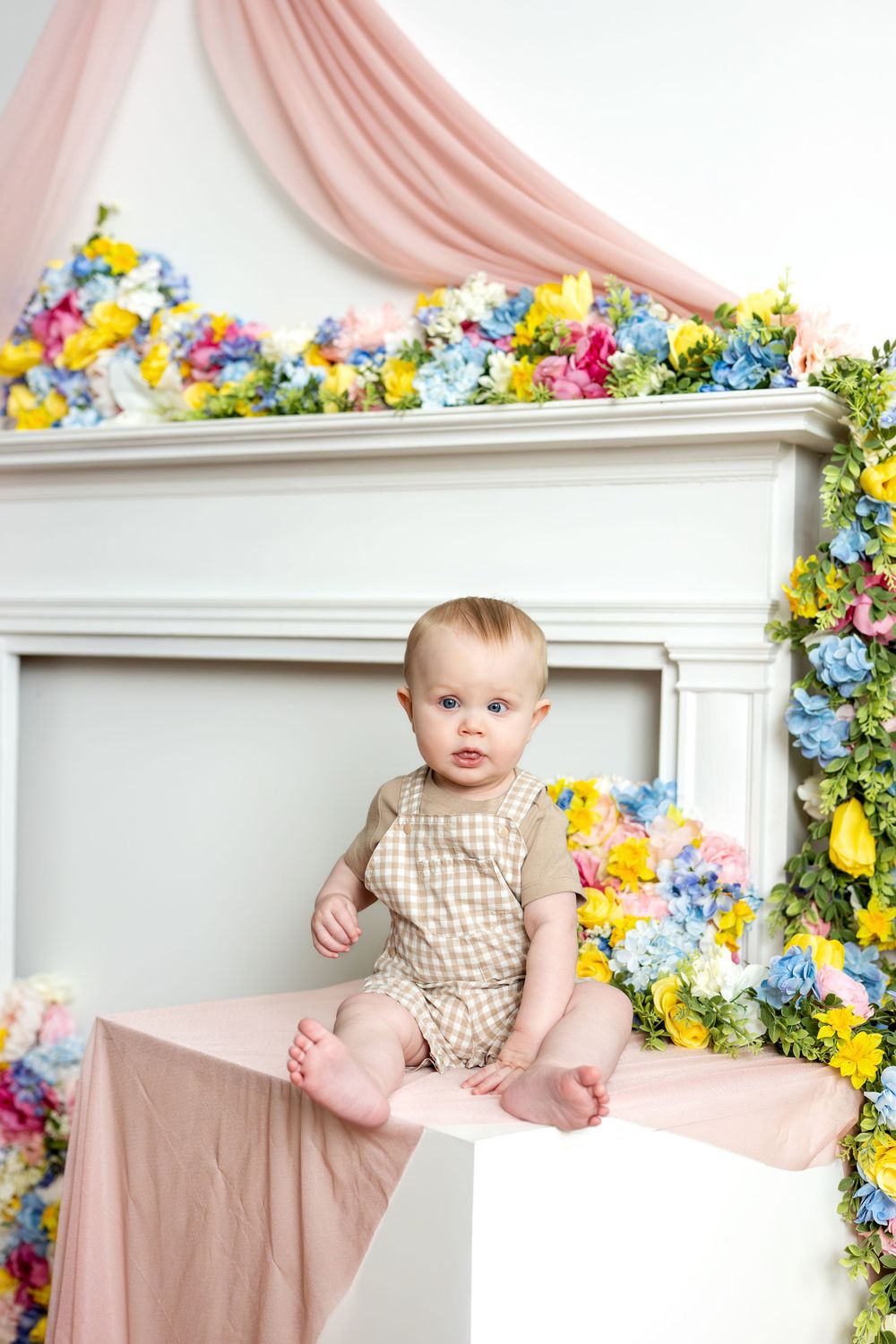 Sweet baby in plaid romper sitting on white cube surrounded by vibrant spring flowers in studio.