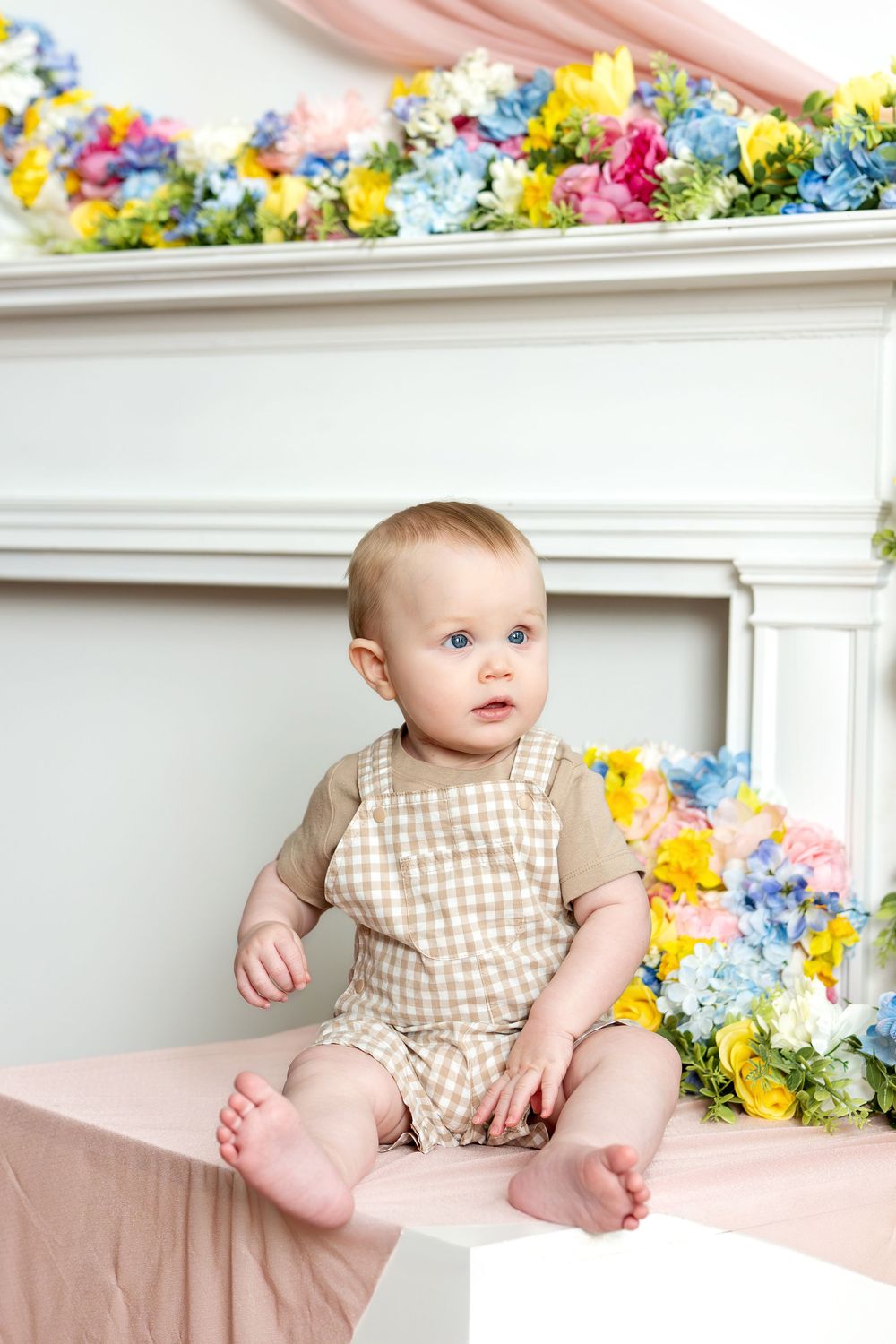 Cute baby with blue eyes sitting barefoot on white cube with colorful spring floral backdrop.