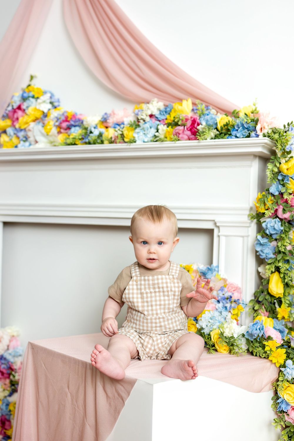 Baby in beige plaid romper sitting barefoot near colorful spring floral arrangement in white studio.