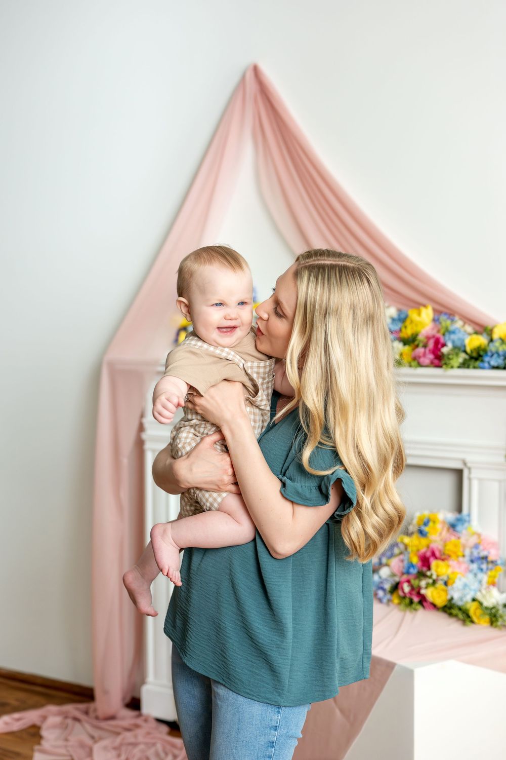Happy blonde mother kissing laughing baby in bright spring studio with colorful floral backdrop.