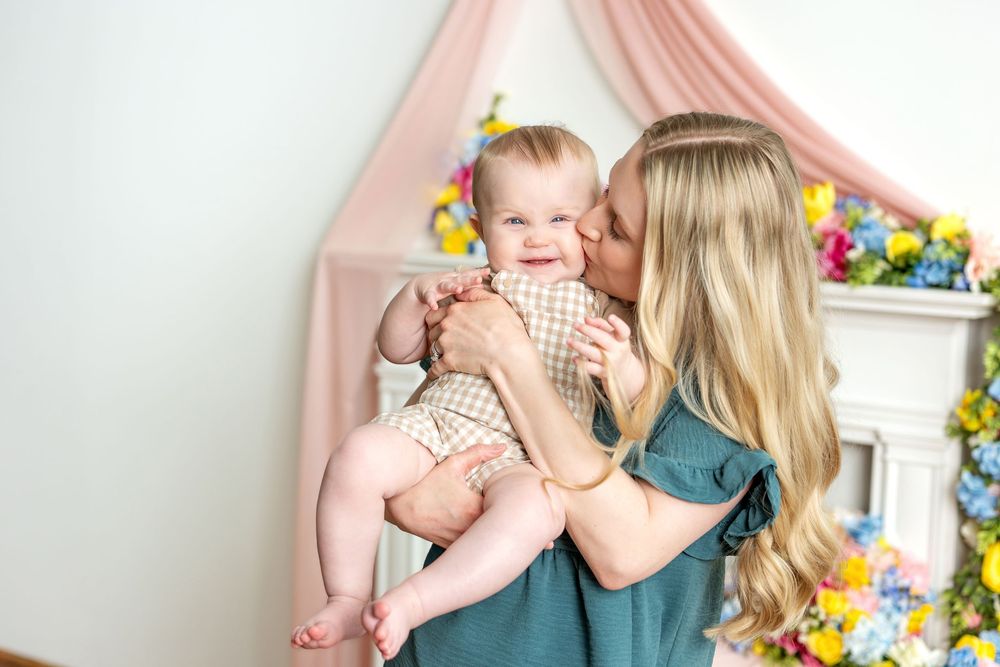 Loving mother kissing her giggling baby in a pastel spring-themed photo studio with flowers.