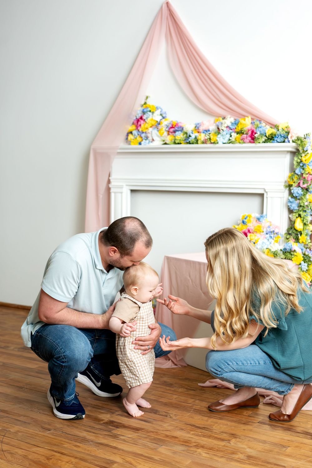 Parents crouching with baby standing between them near floral fireplace.