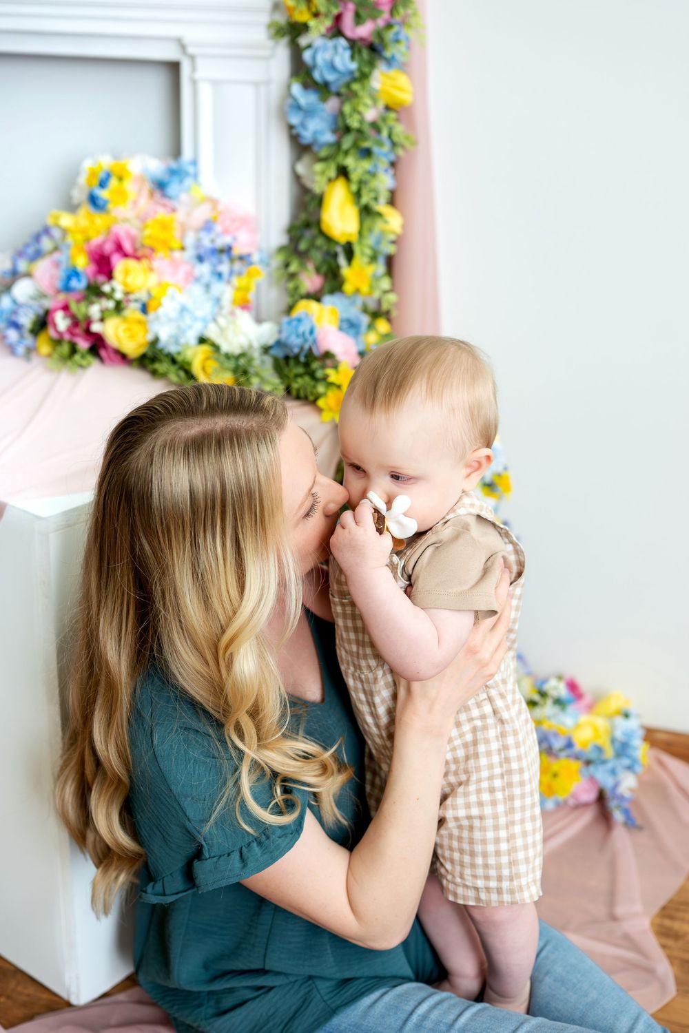 Mother kissing smiling baby near colorful spring floral arrangement.