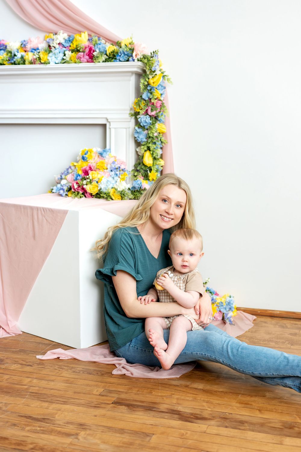 Smiling mother sitting with baby on lap near floral fireplace decor.
