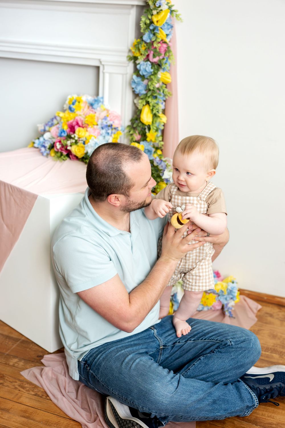 Father sitting cross-legged on floor playing with smiling baby in floral spring photo studio.