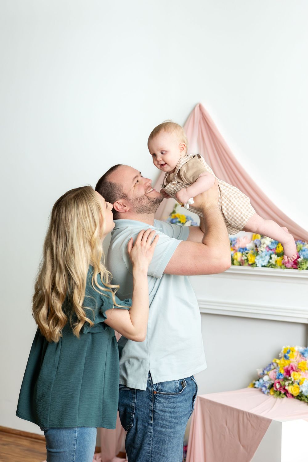 Happy family with mother and father lifting laughing baby in colorful floral spring studio.