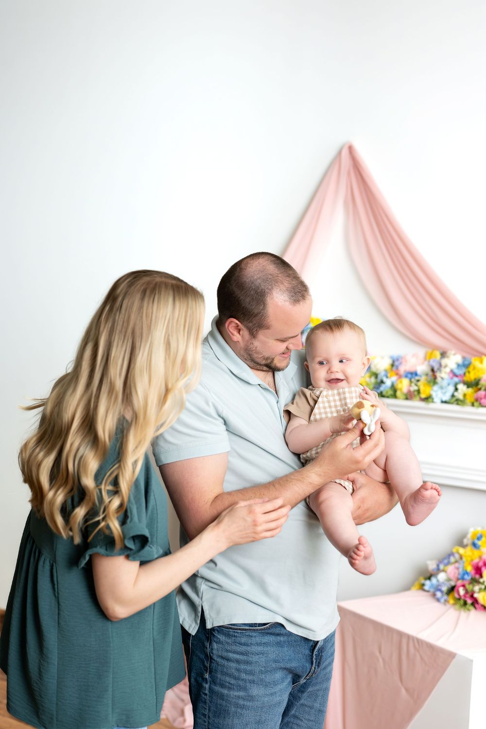 Young family with mother and father holding happy baby in bright spring floral studio.