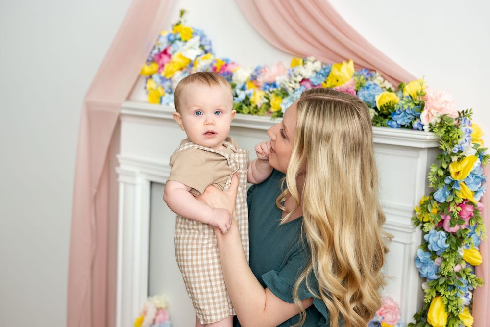 Blonde mother kissing infant baby held in arms near spring floral decorated fireplace.