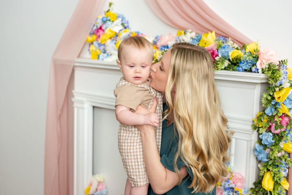 Blonde mother kissing her baby in a floral spring photo studio with colorful flowers and pink draping.