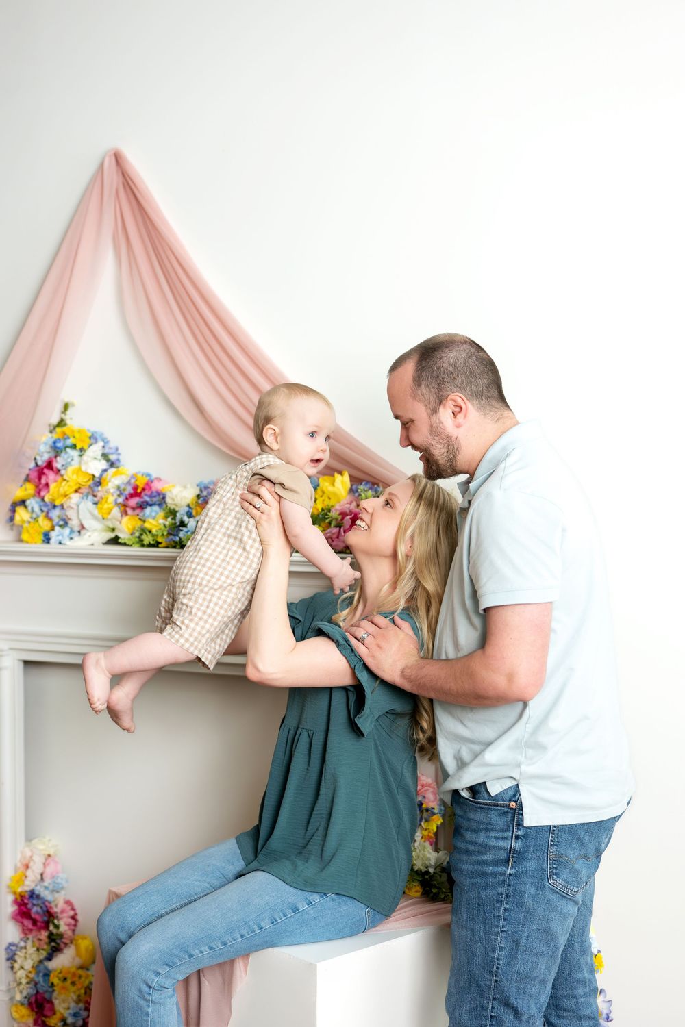 Happy parents lifting laughing baby together near colorful spring floral fireplace decoration.