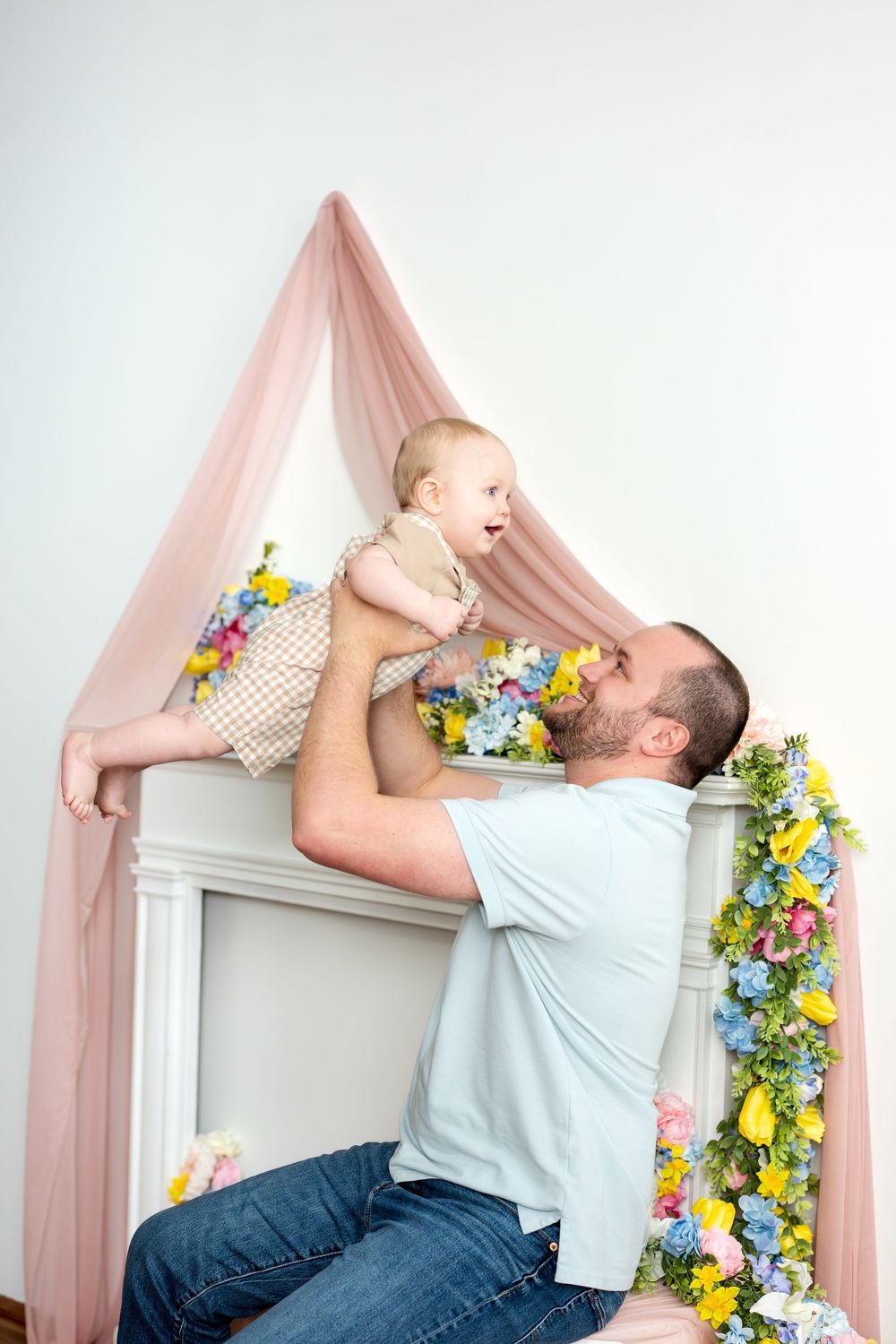 Father lifting happy baby up near spring floral decorated fireplace with pink draped backdrop.