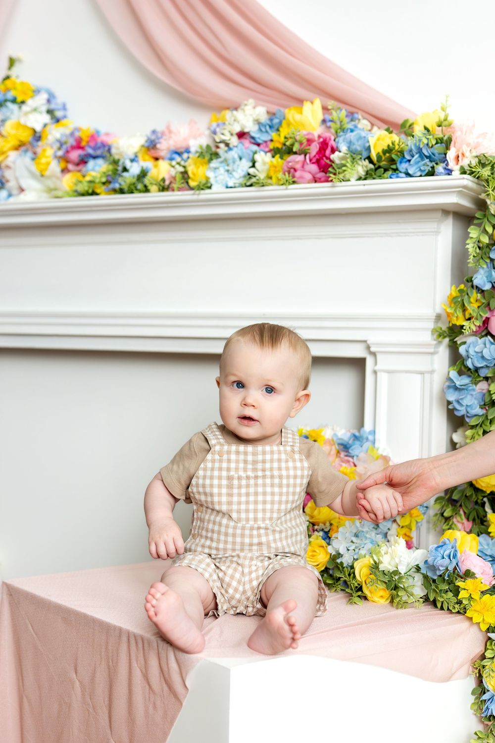 Adorable blue-eyed baby sitting on white cube surrounded by colorful spring flowers in photo studio.
