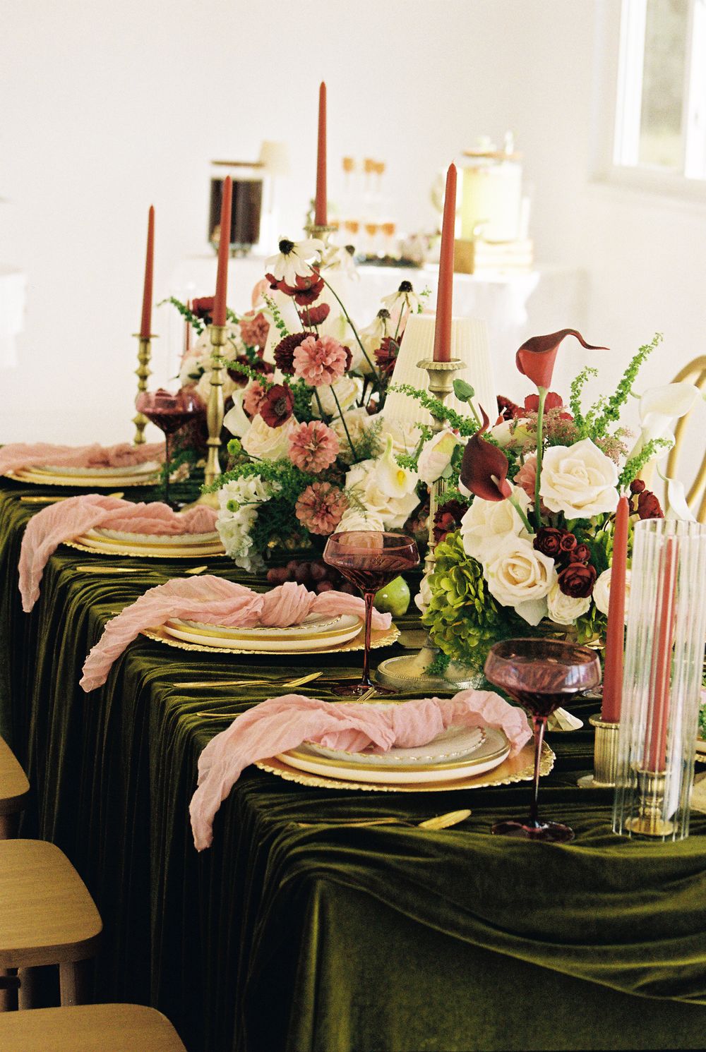 Elegant wedding tablescape with green velvet tablecloth, pink napkins, gold chargers, red candles, and lush floral centerpieces.