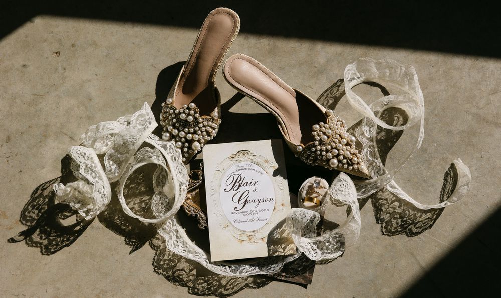 Elegant wedding flat lay with gold heels, lace, pearls, and calligraphy invitation on dark background.