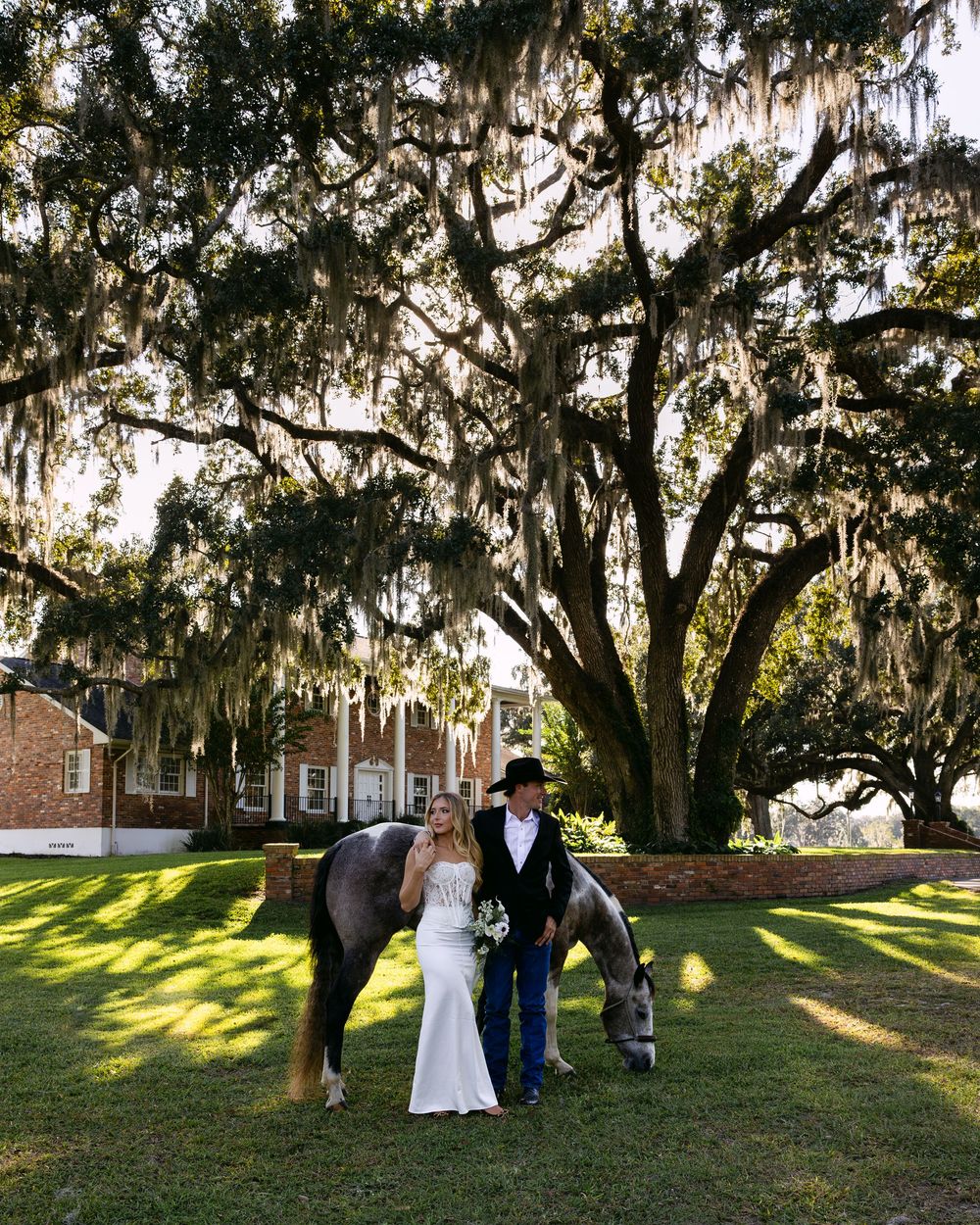 Bride and groom stand with a grazing horse under sprawling Spanish moss oak trees at outdoor wedding venue.