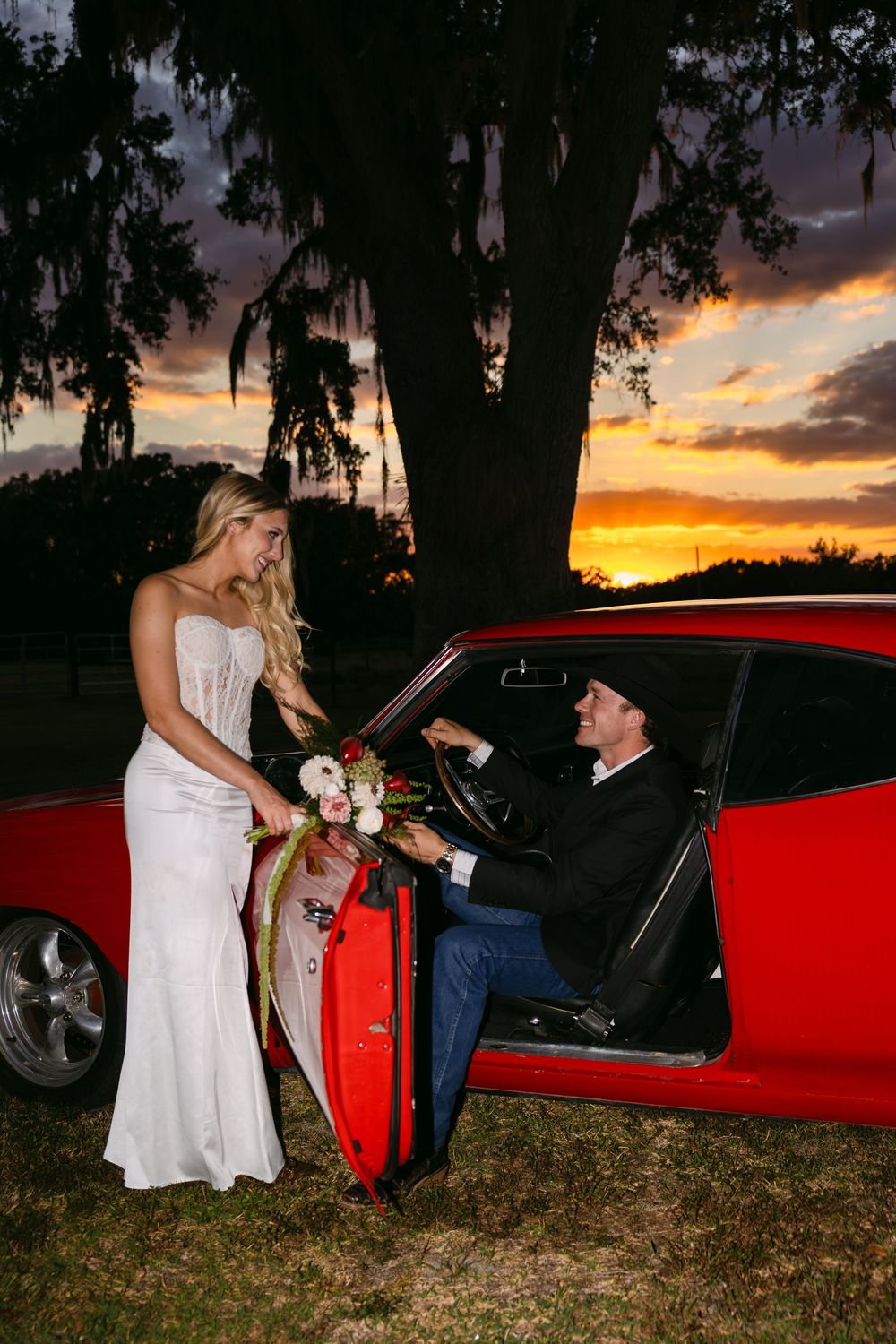 Bride in white gown holds bouquet beside red sports car at sunset, as groom sits in driver seat smiling.