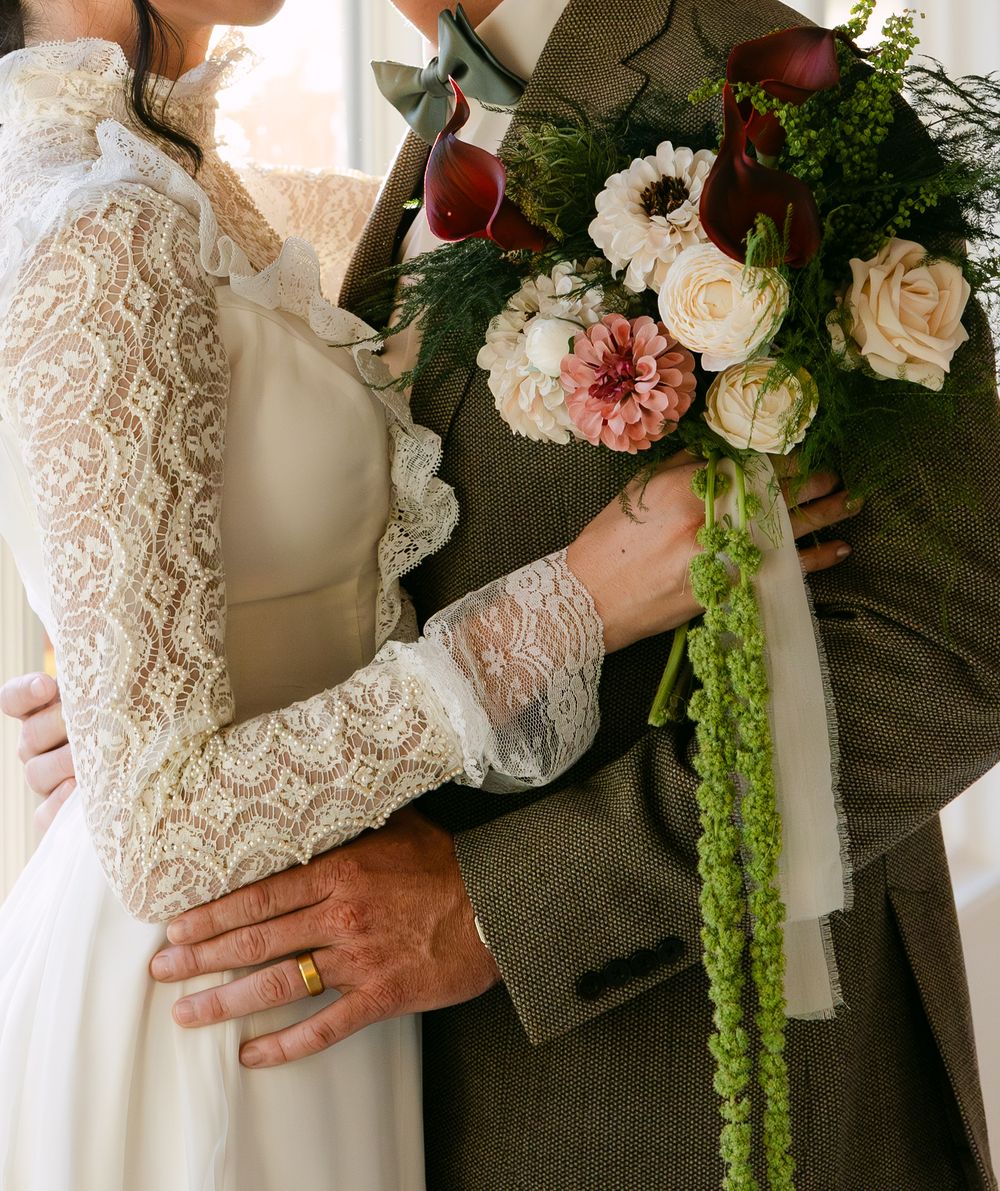 Bride in lace dress holds bouquet of roses and dahlias while groom in tweed suit embraces her at wedding.