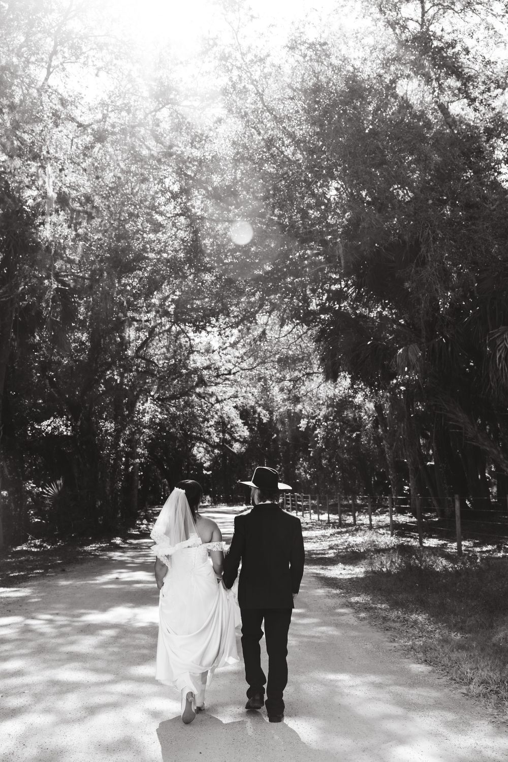 Black and white photo of bride and groom walking hand in hand down a tree-lined path on their wedding day.
