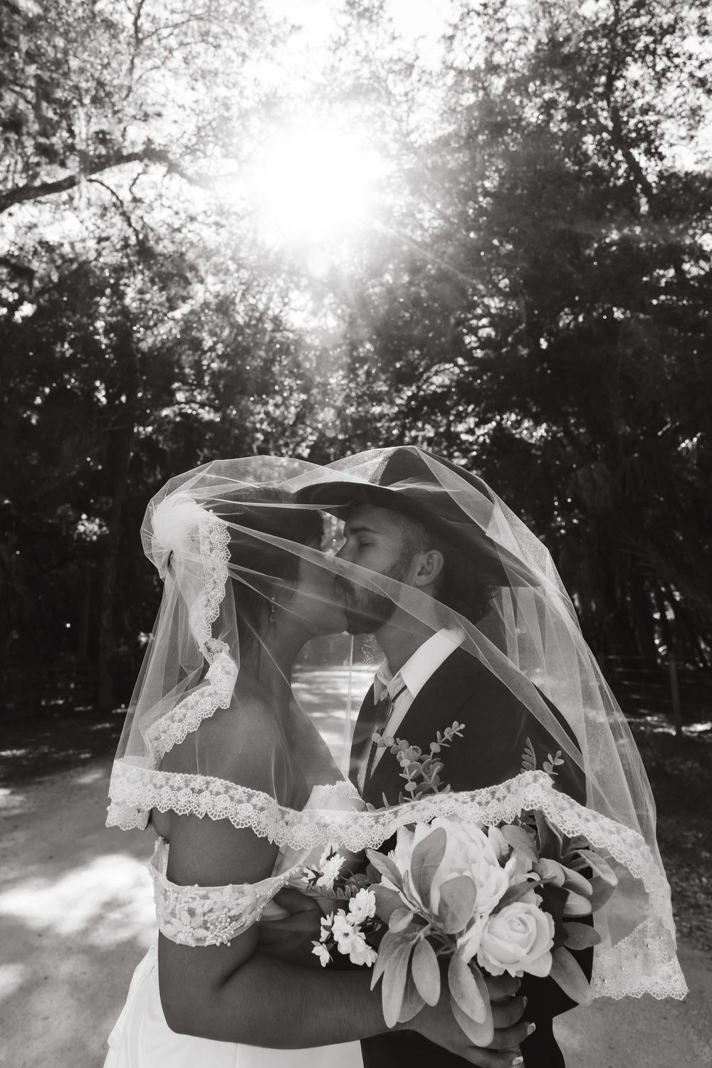 Black and white photo of bride and groom kissing under wedding veil outdoors with sunlight filtering through trees.