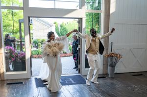 Joyful bride and groom making grand entrance into rustic wedding venue with feather bouquet and gold attire.