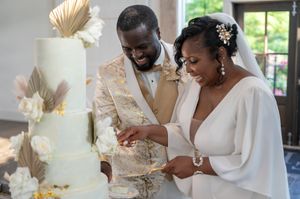 Happy couple cutting elegant white wedding cake with gold details and floral decorations at their reception.