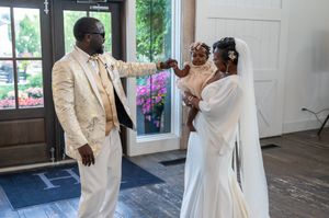 Groom in gold suit touches baby held by bride in white gown and veil at wedding venue.