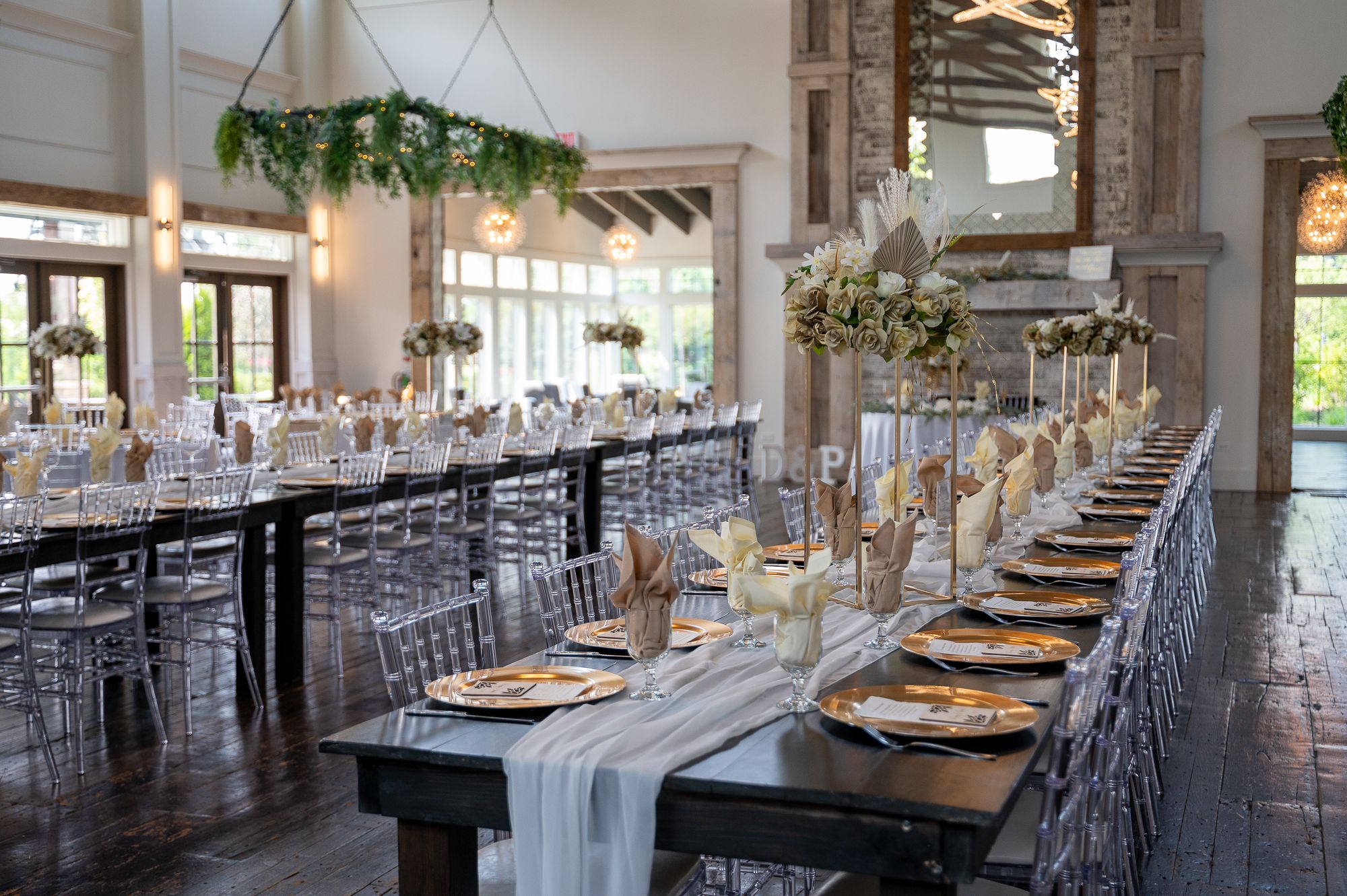 Elegant wedding reception hall with long farm tables, ghost chairs, floral centerpieces, and greenery chandelier.