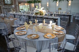 Elegant wedding reception hall with round tables, silver chiavari chairs, gold charger plates, and floral centerpieces.