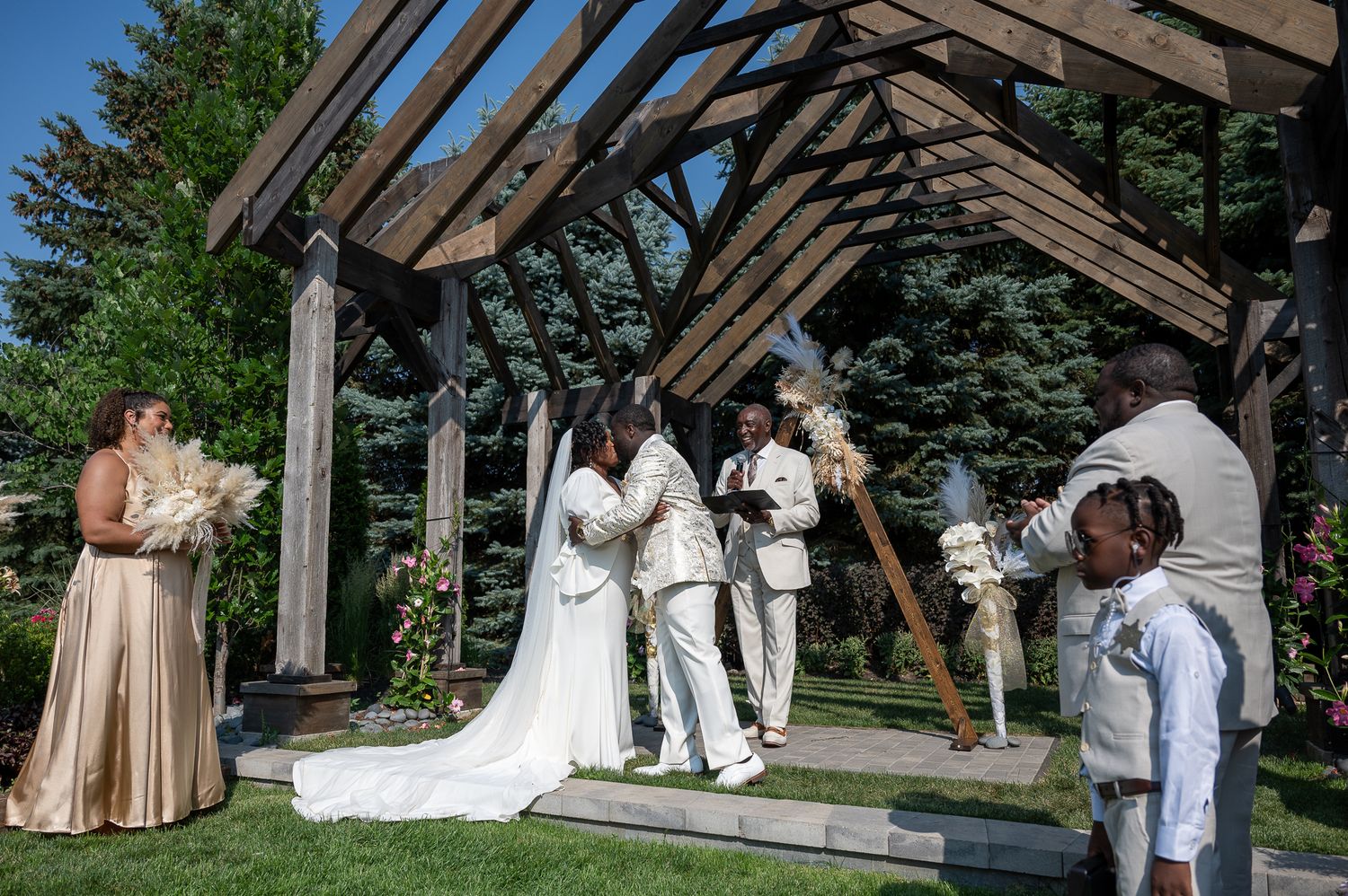 Outdoor wedding ceremony under wooden pergola with bride, groom, officiant, and guests on sunny day.