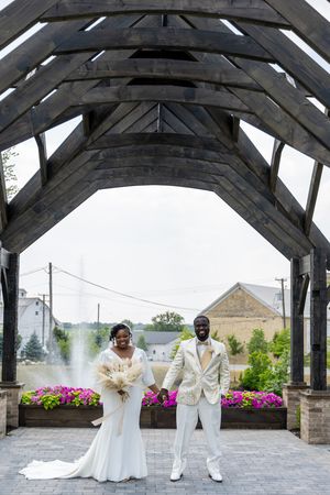 Bride and groom stand under rustic wooden arch at outdoor wedding venue with colorful flower beds and fountain backdrop.