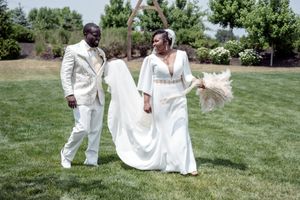 A Black couple in elegant cream wedding attire walks on green grass, bride holding pampas grass bouquet.