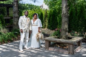 Elegant couple in white and gold wedding attire posing in a lush garden with a rustic wooden bench.