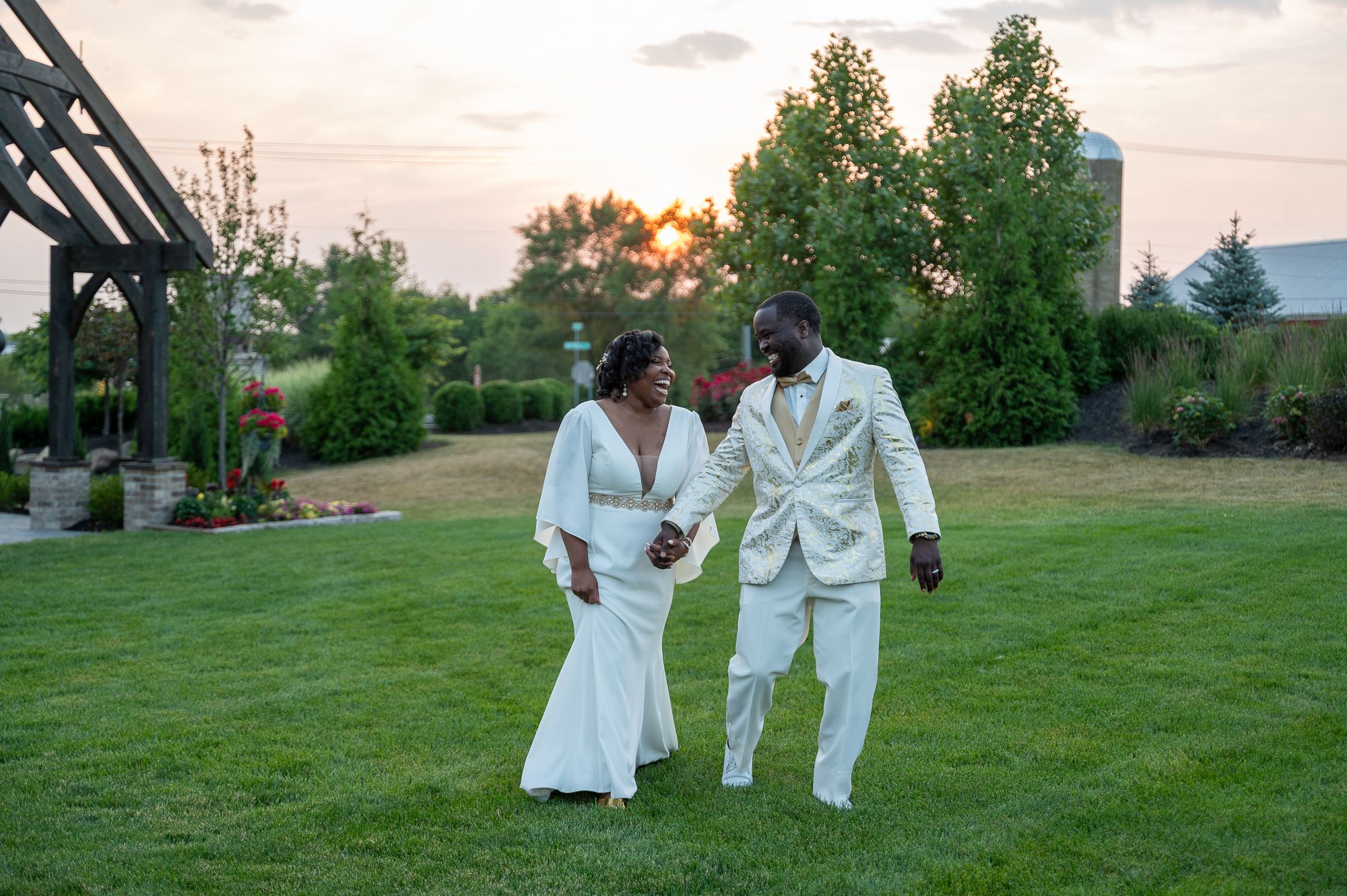 Newlywed couple in white attire holding hands and walking on green lawn at sunset during outdoor wedding celebration.