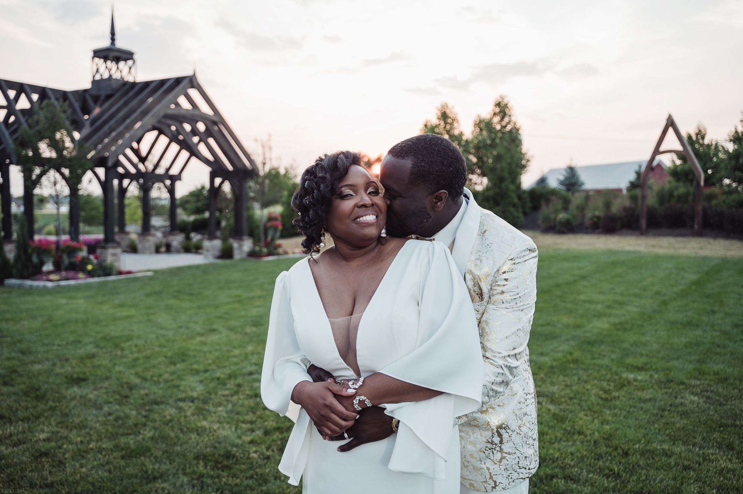 A joyful couple in elegant wedding attire share a romantic moment on a lush green lawn at sunset.