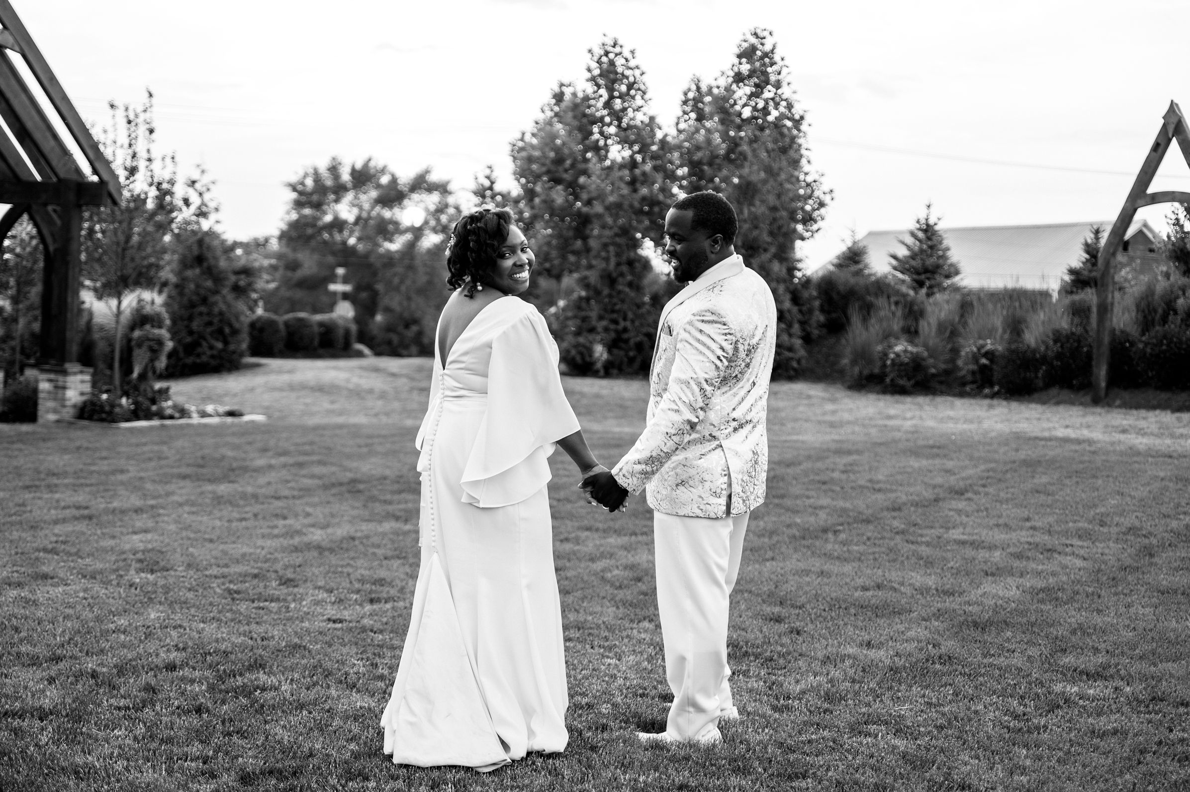 Black and white photo of a couple holding hands on a lawn, dressed in white wedding attire, surrounded by trees.