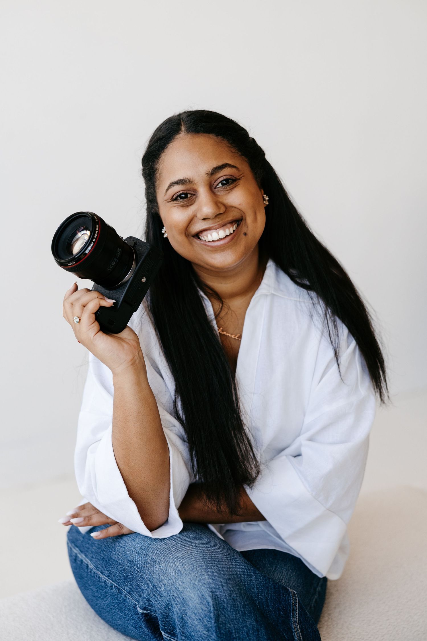 Smiling female photographer holding DSLR camera sitting cross-legged against white background.