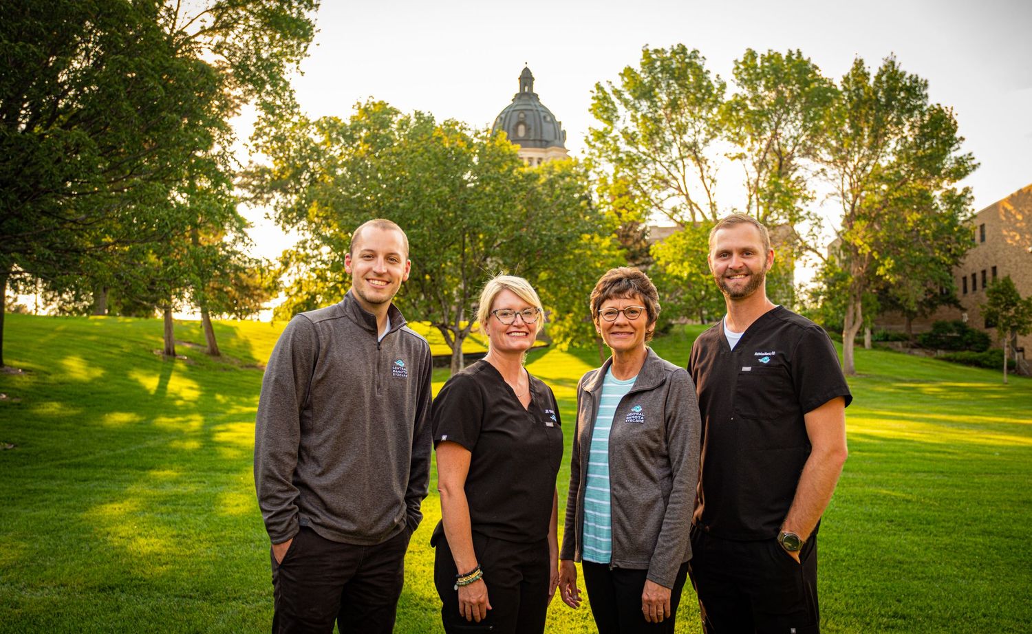 Four people stand together on a green lawn with a domed building visible in the background at sunset.