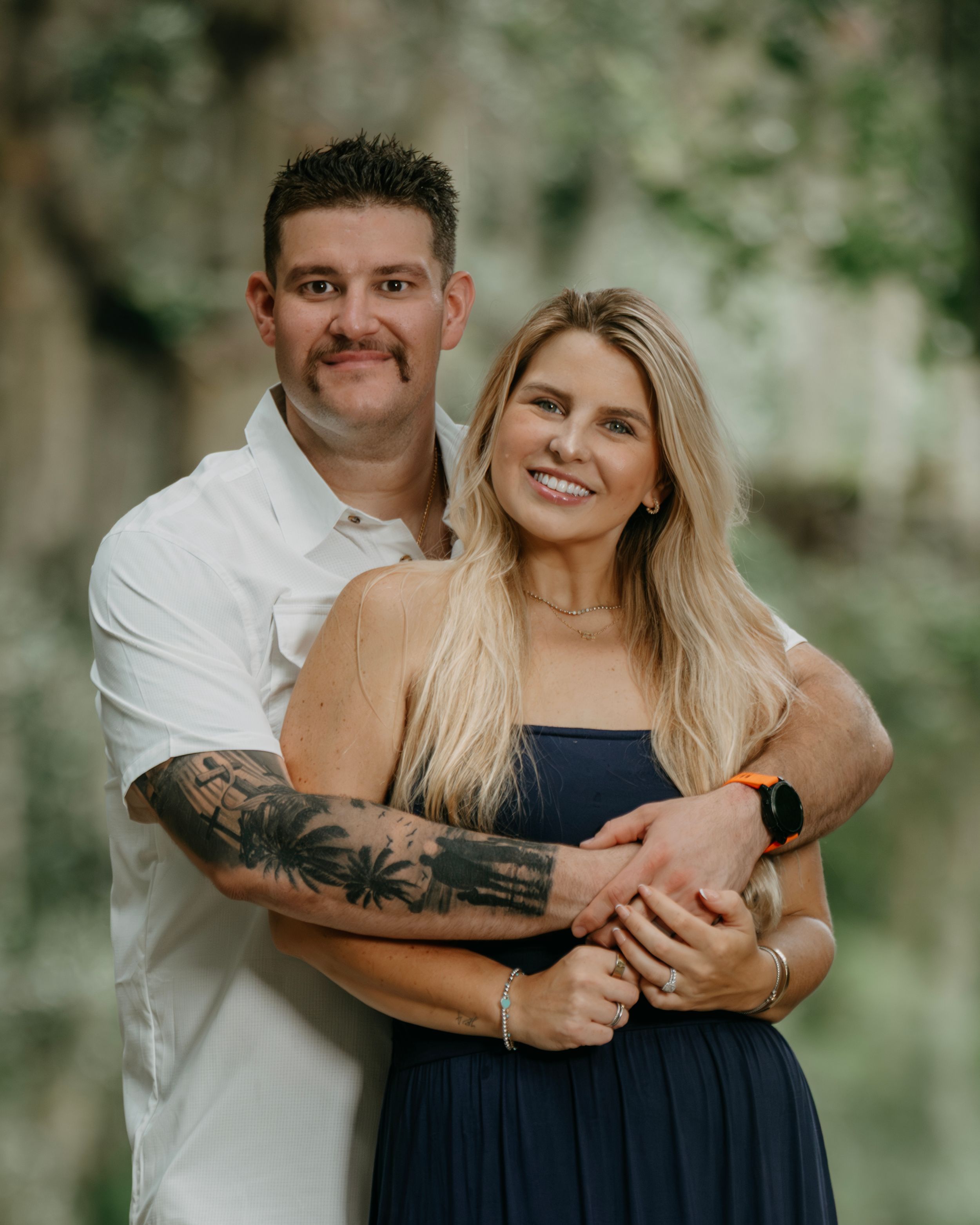 A smiling couple poses outdoors, man in white shirt with tattoos embracing a blonde woman in a navy dress.