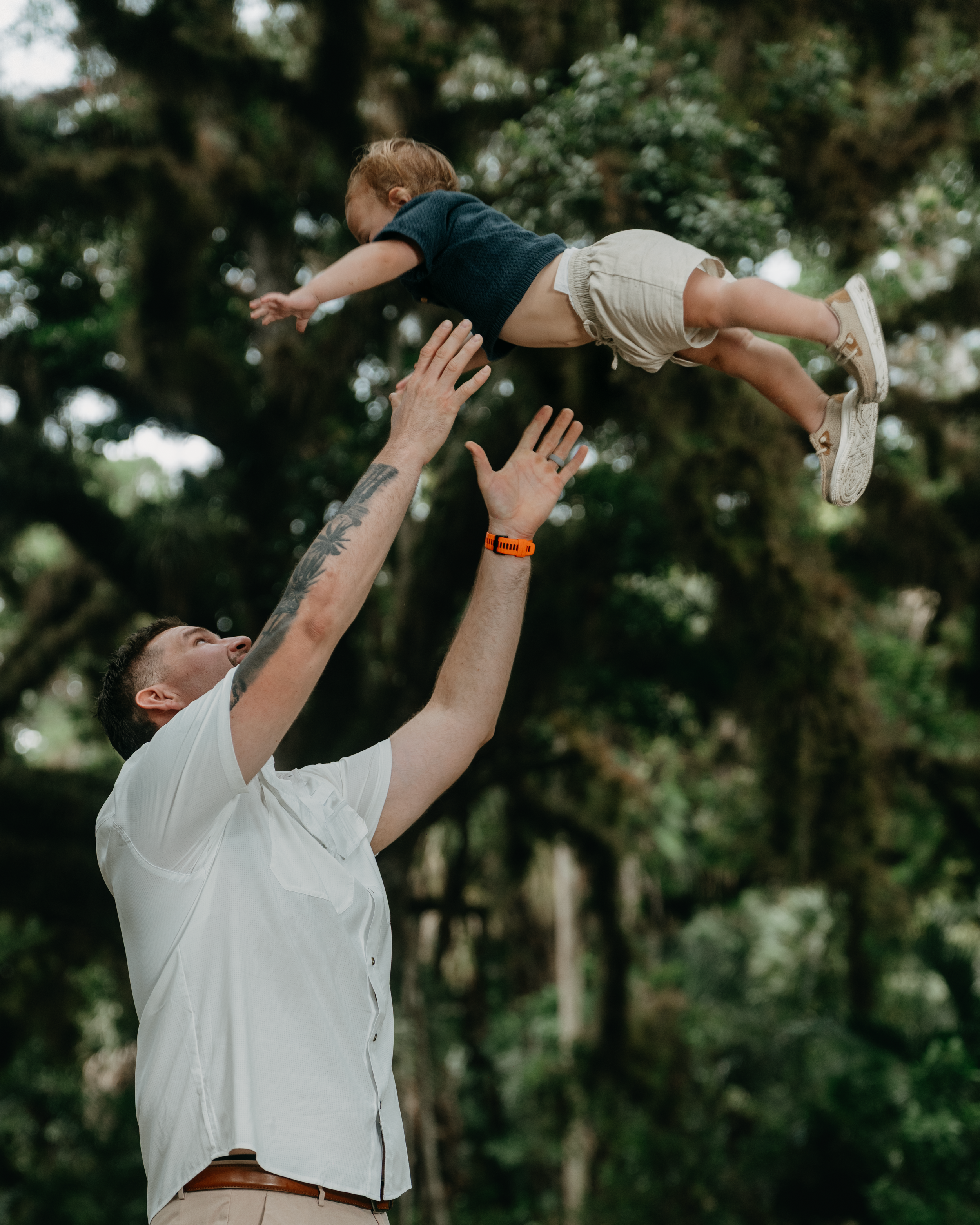 A father joyfully tosses his young child into the air outdoors, surrounded by lush green trees.