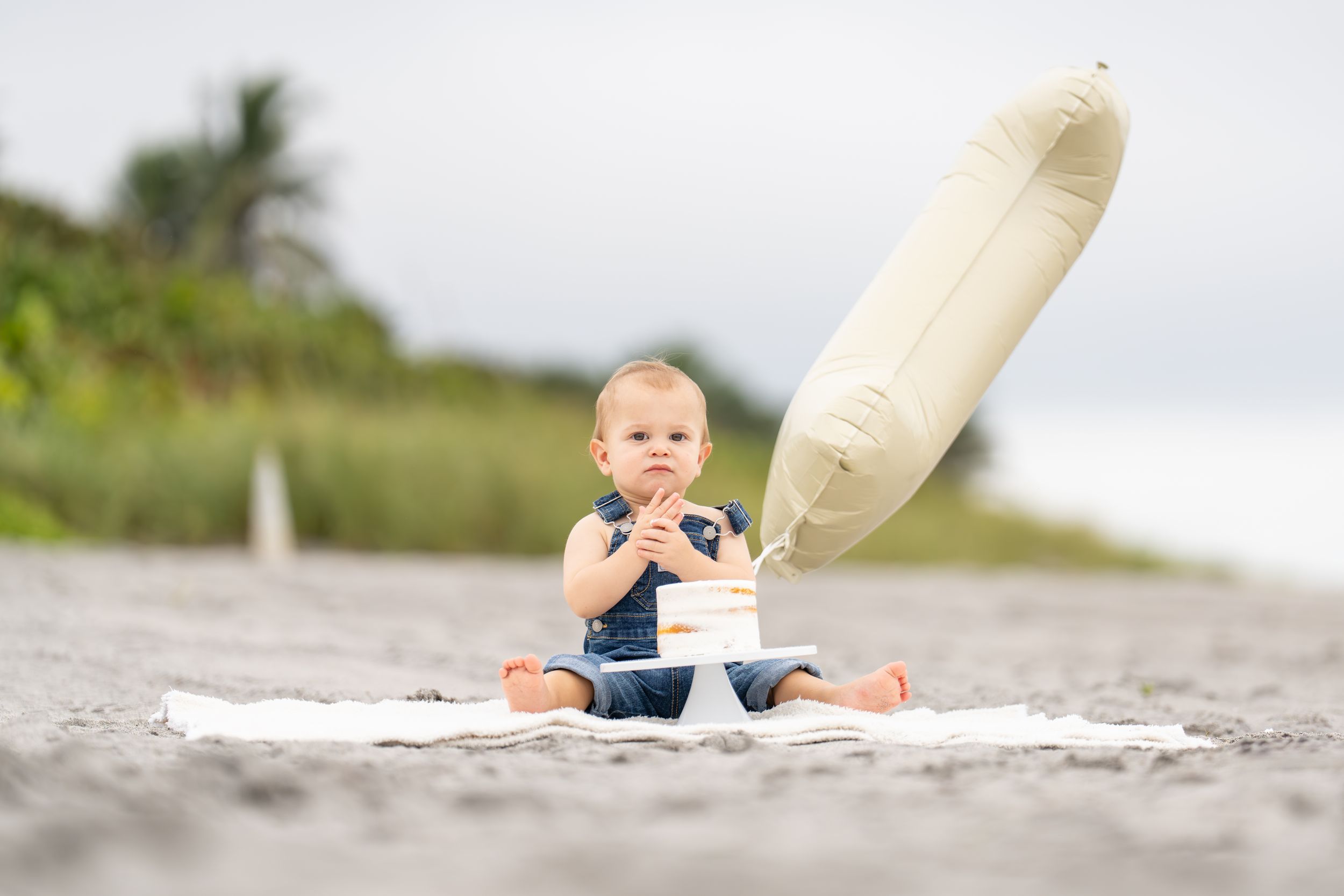 A small surfer sits on a sandy beach with a large white surfboard beside them on a cloudy day.