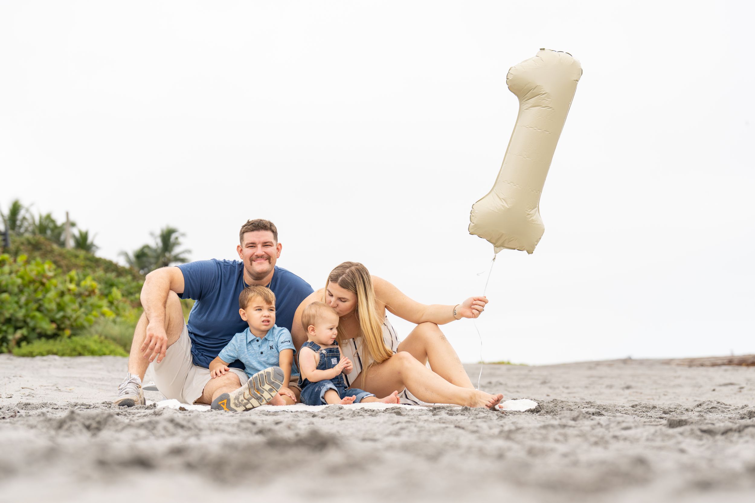 Family relaxing together on sandy beach with floating bone-shaped pool toy in the air.