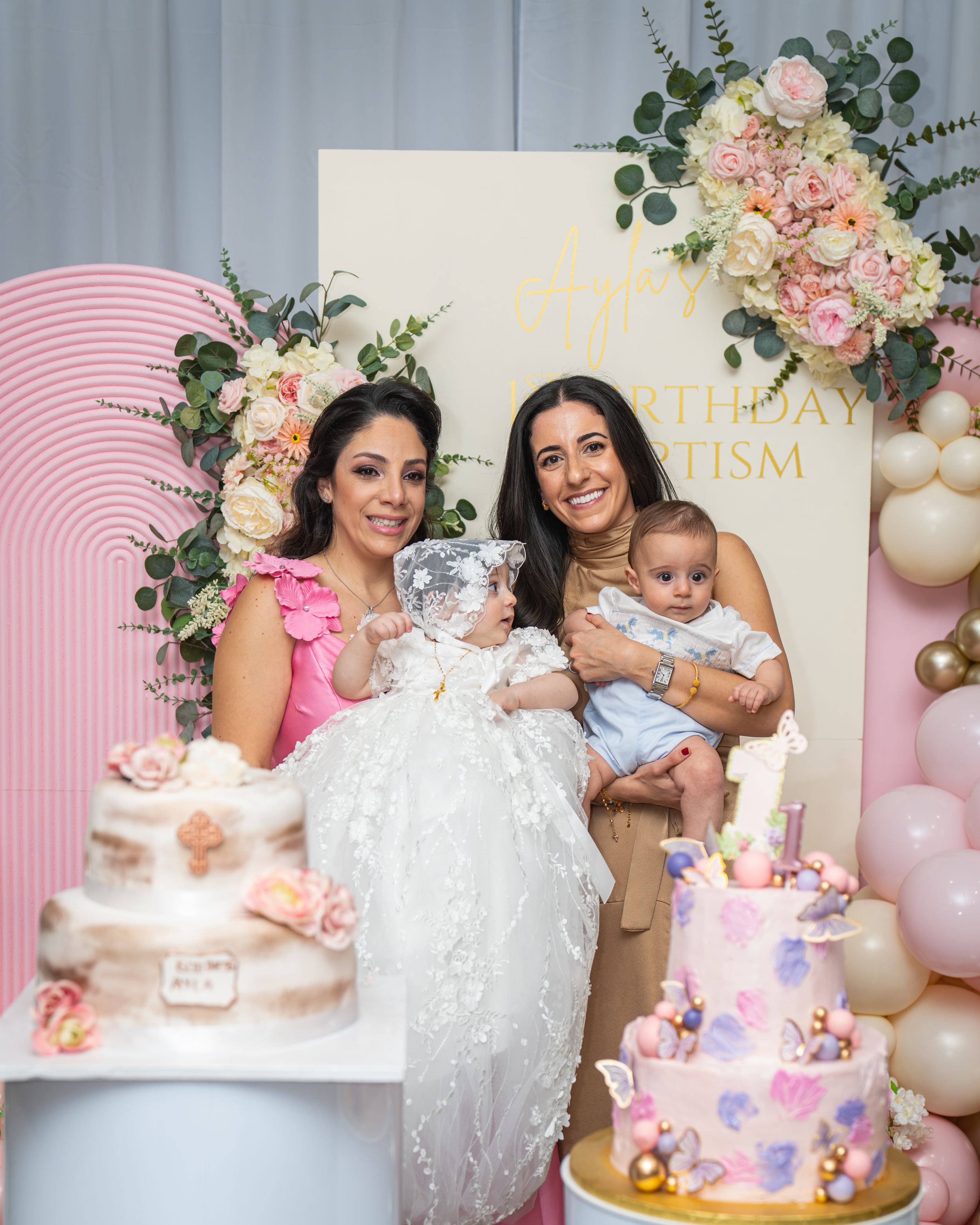 A baby's first birthday party setup with pink balloons, floral decorations, and a white cake on display.