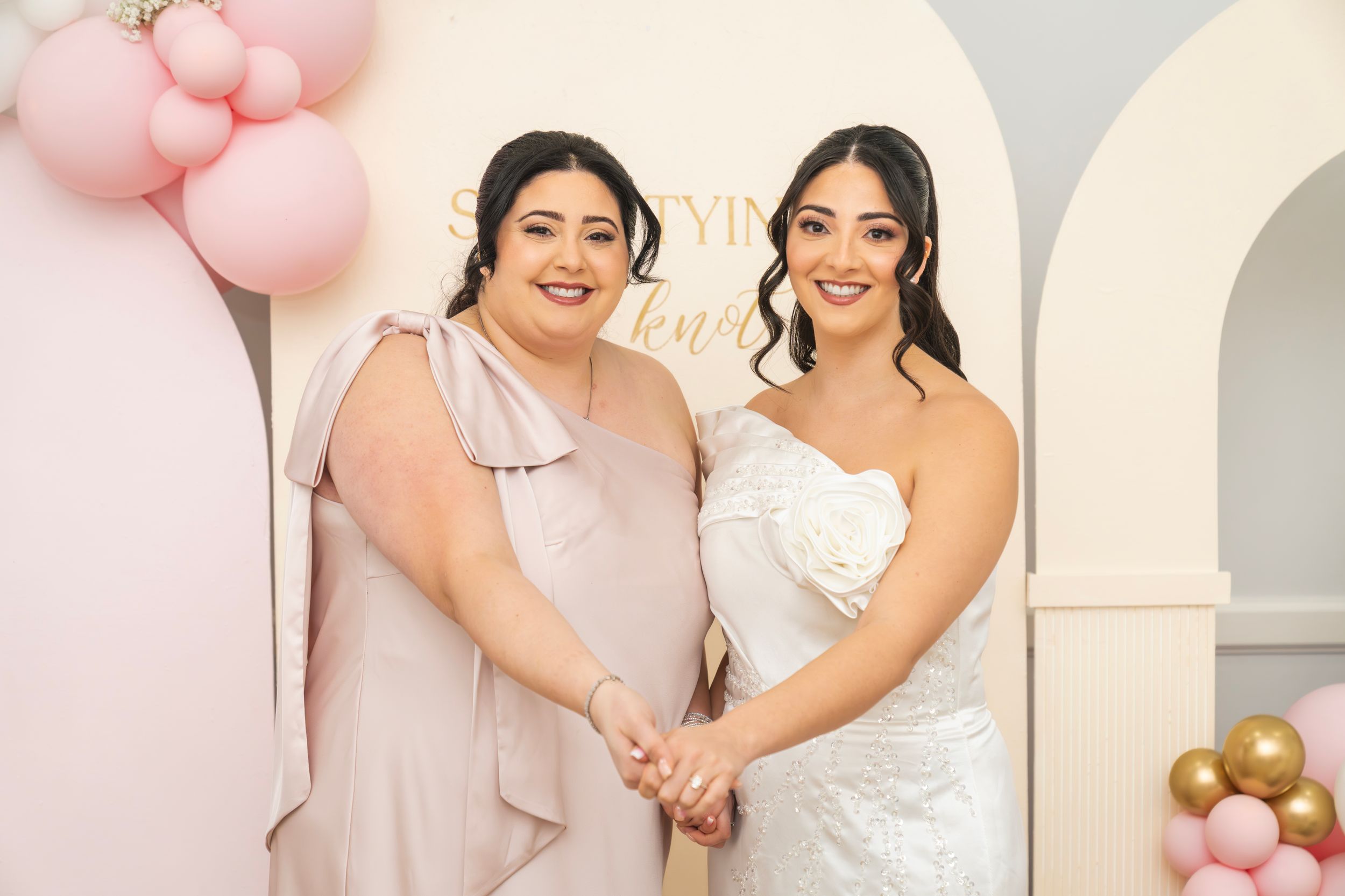 Wedding party poses together in front of white arches with pink balloons and decorative floral arrangements.