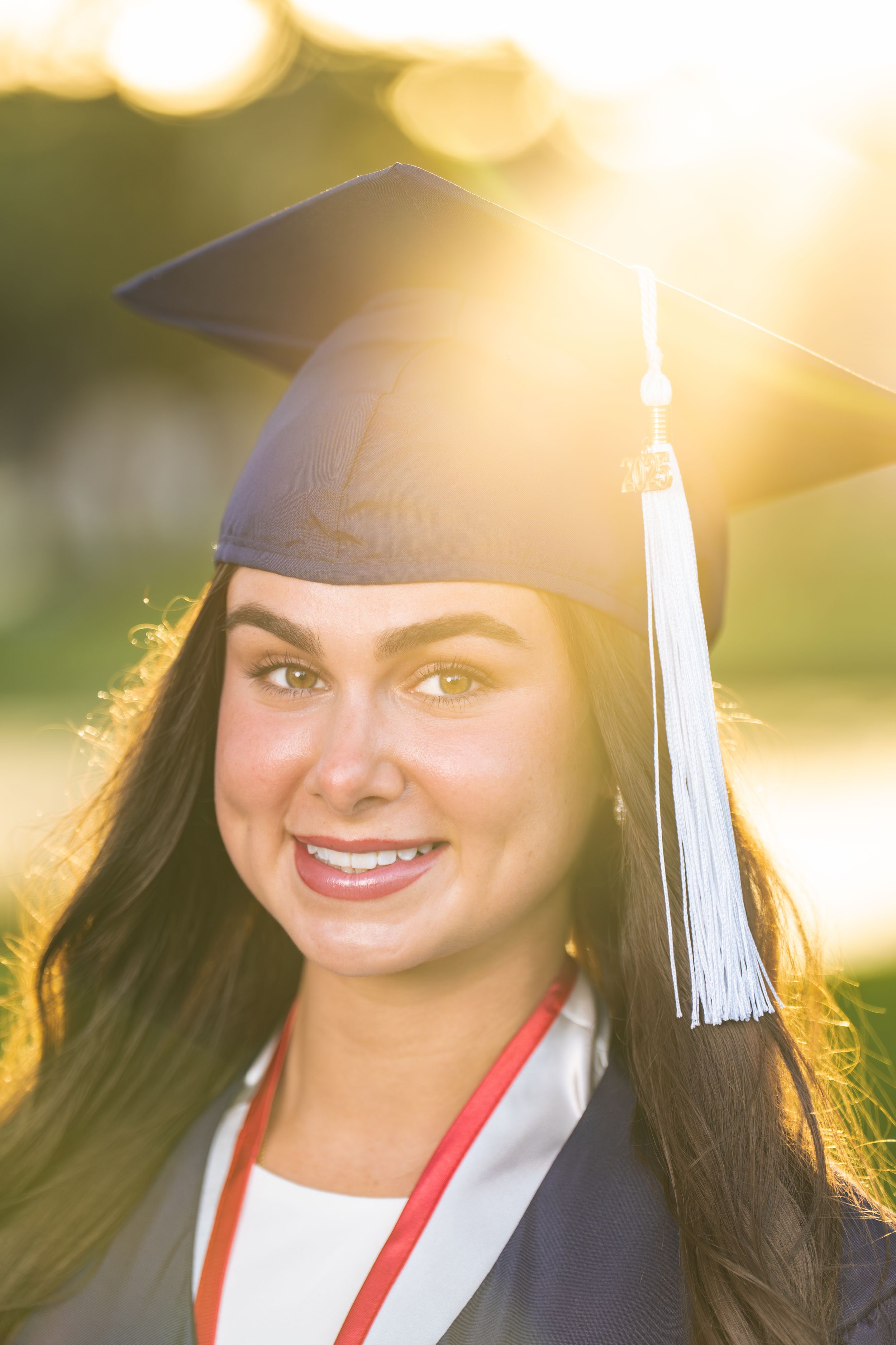 Graduate in cap and gown smiling during golden hour sunset with white tassel and red graduation stole.