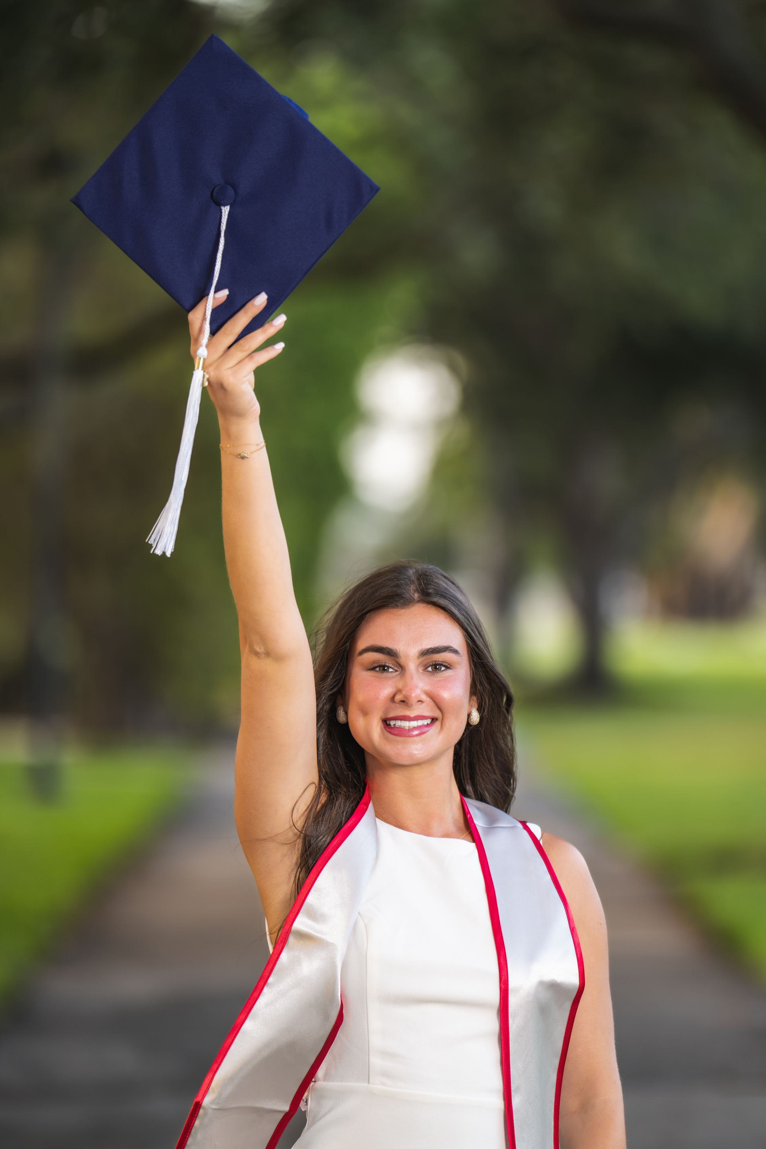 Graduate in white dress and navy cap holds up graduation cap while celebrating outdoors on tree-lined path.