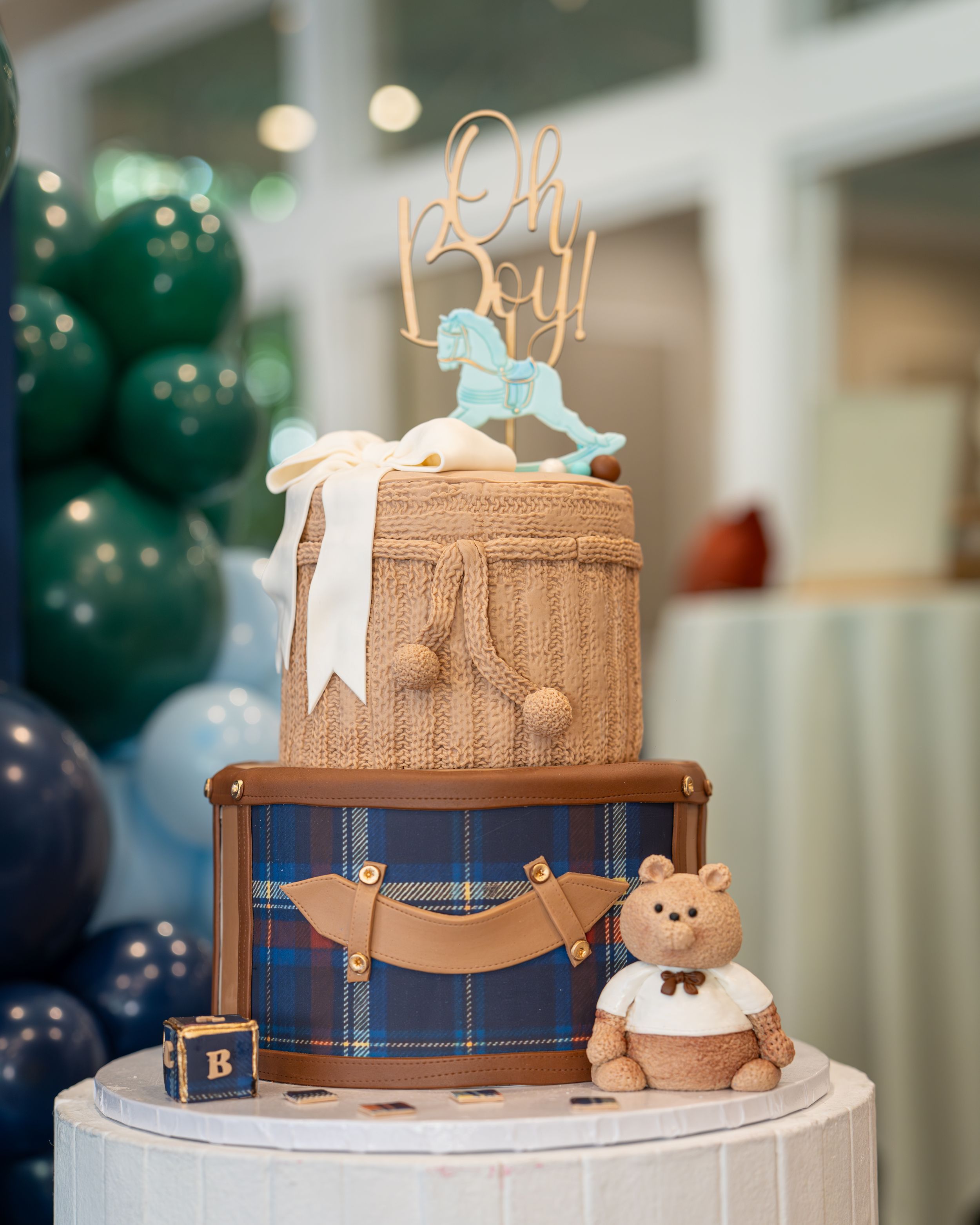 Two-tier baby shower cake decorated in brown and beige with a teddy bear topper and blue bow accents on a white stand.