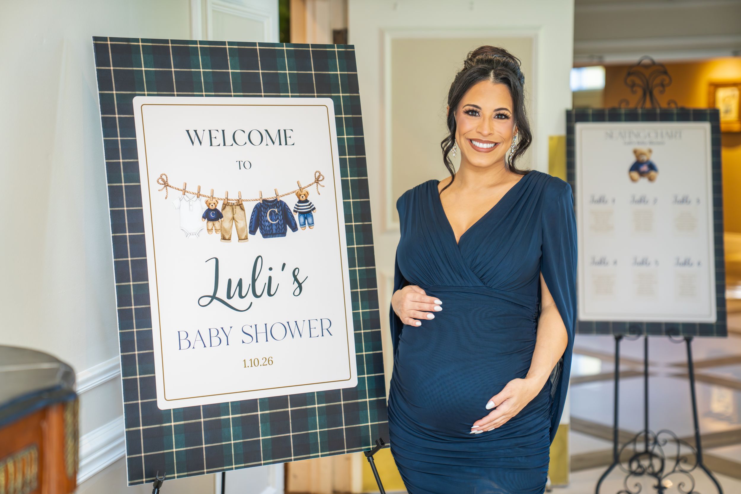 A navy blue baby shower welcome sign with clothing illustrations stands next to someone in a matching navy dress.