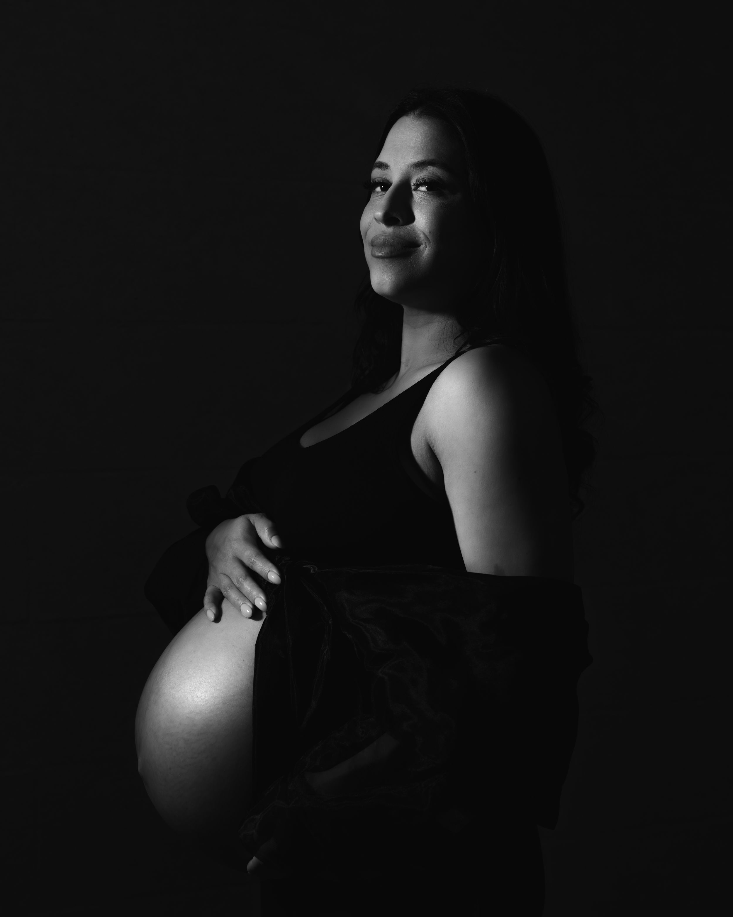 A dramatic black and white maternity portrait with artistic lighting showcasing an expectant mother in a dark dress.